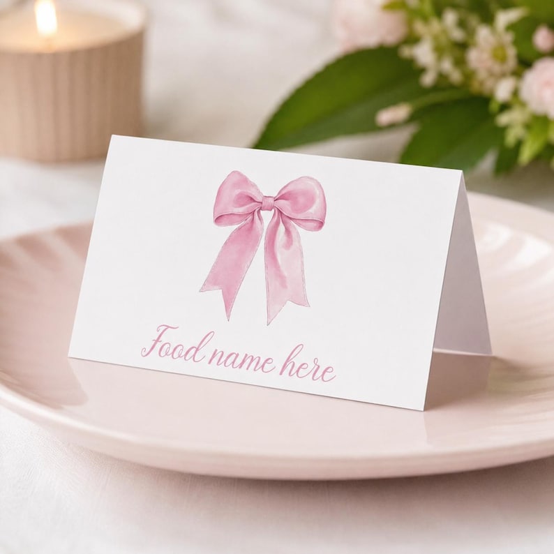 May include: A white folded place card featuring a soft pink watercolor illustration of a ribbon bow centered at the top. Below the bow, the text "Food name here" is printed in a light pink cursive script font on the front of the card.