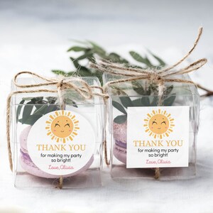 May include: Two clear square favor boxes tied with twine, each containing a light purple macaron and green leaves. A label on each box features a yellow sun illustration and the text: THANK YOU for making my party so bright! Love, Olivia.