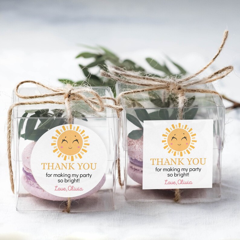 May include: Two clear square favor boxes tied with twine, each containing a light purple macaron and green leaves. A label on each box features a yellow sun illustration and the text: THANK YOU for making my party so bright! Love, Olivia.