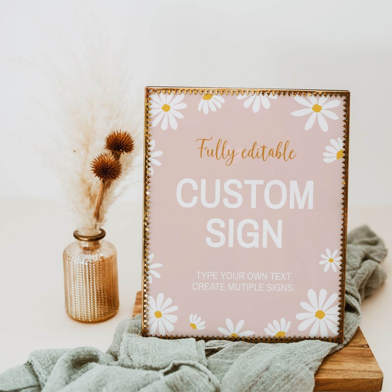 May include: A rectangular sign in a gold metal frame with a light pink background featuring white daisy illustrations. The text reads Fully editable CUSTOM SIGN TYPE YOUR OWN TEXT CREATE MUTIPLE SIGNS. The sign is displayed next to a textured glass vase.