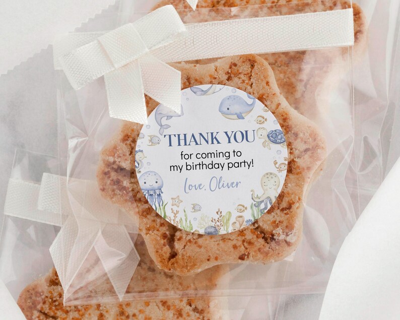 May include: A star-shaped cookie in a clear cellophane bag, tied with a white ribbon and bow. A circular sticker reads "THANK YOU for coming to my birthday party! Love, Oliver" with sea creature illustrations.