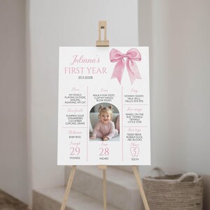 May include: A pink and white first year milestone poster with a watercolor bow at the top. The poster includes information about the baby's first year, including their weight, height, favorite foods, and activities. The poster also includes a photo of the baby.