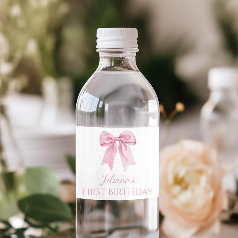 Puede incluir: Una botella de agua de pl&aacute;stico transparente con tapa blanca y una etiqueta rectangular. La etiqueta presenta una ilustraci&oacute;n de un lazo de acuarela rosa sobre el texto "Juliana's FIRST BIRTHDAY" impreso en una fuente serif rosa claro.