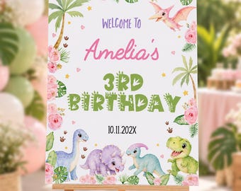May include: A rectangular birthday welcome sign featuring a white background with pink floral and green tropical leaf borders. The text reads Welcome to Amelia's 3rd Birthday 10.11.202X, accompanied by illustrations of a pterodactyl and three dinosaurs.