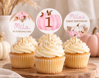 May include: Three cupcakes topped with round paper toppers on wooden picks. The toppers feature pink pumpkins, floral accents, and text reading Mila IS TURNING ONE, the number 1, and Mila's FIRST BIRTHDAY in brown and pink script and block fonts.