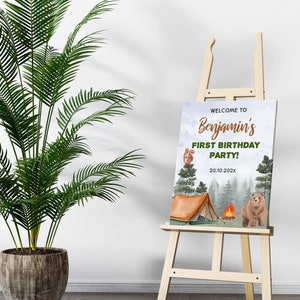 Op de afbeelding: Een rechthoekig bord op een houten ezel met een kampeerthema, een bruine beer, een eekhoorn, een tent en een kampvuur. De tekst luidt: WELCOME TO Benjamin's FIRST BIRTHDAY PARTY! 20.10.202X. De achtergrond toont een bos met groene bomen.