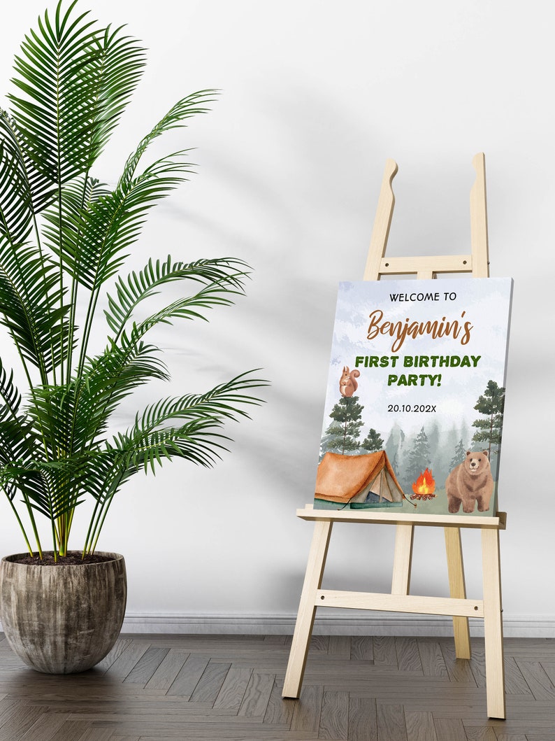 Op de afbeelding: Een rechthoekig bord op een houten ezel met een kampeerthema, een bruine beer, een eekhoorn, een tent en een kampvuur. De tekst luidt: WELCOME TO Benjamin's FIRST BIRTHDAY PARTY! 20.10.202X. De achtergrond toont een bos met groene bomen.