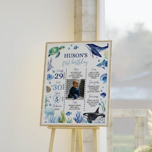 May include: A blue and white first birthday milestone poster with a photo of a baby boy. The poster features a sea-themed design with fish, whales, and a turtle. The poster lists the baby's age, weight, height, and other milestones.