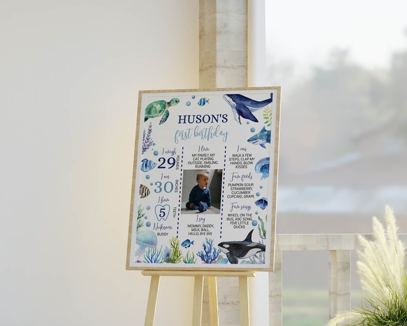 May include: A blue and white first birthday milestone poster with a photo of a baby boy. The poster features a sea-themed design with fish, whales, and a turtle. The poster lists the baby's age, weight, height, and other milestones.