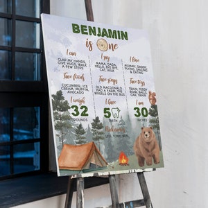 May include: A rectangular milestone birthday sign for a one-year-old named Benjamin, featuring a forest theme with a brown bear, a tent, and a campfire. The text details personal milestones, including weight of 32 pounds and height of 30 inches.