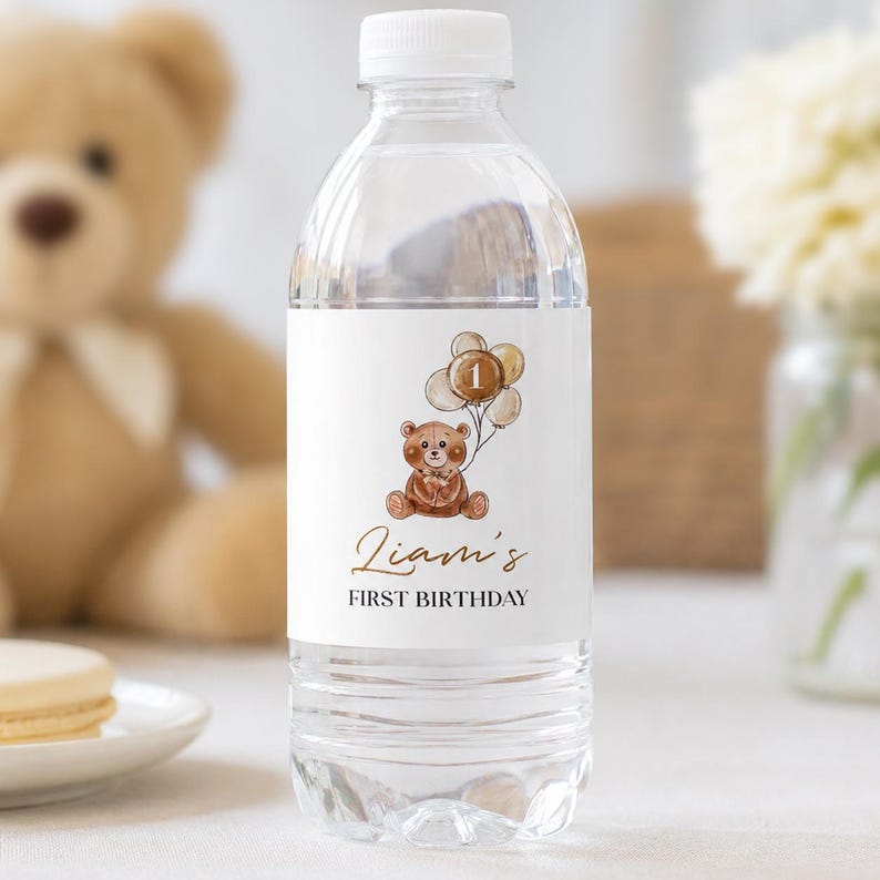 May include: A clear plastic water bottle with a white label featuring a brown teddy bear illustration holding a bunch of balloons with the number one inside one balloon. The text reads Liam's FIRST BIRTHDAY in brown and black script and print.