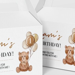 May include: Two white cardboard gable boxes featuring a watercolour illustration of a brown teddy bear holding a cluster of balloons with the number one on the centre balloon. The text reads Liam's 1st BIRTHDAY! THANK YOU FOR CELEBRATING WITH ME!