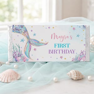 Op de afbeelding: Een rechthoekige wikkel voor een chocoladereep met een illustratie van een zeemeerminstaart, koraal, schelpen en kleine sterretjes. De tekst luidt Mayra's FIRST BIRTHDAY in roze, blauwgroene en paarse letters op een witte achtergrond met gouden stippen.