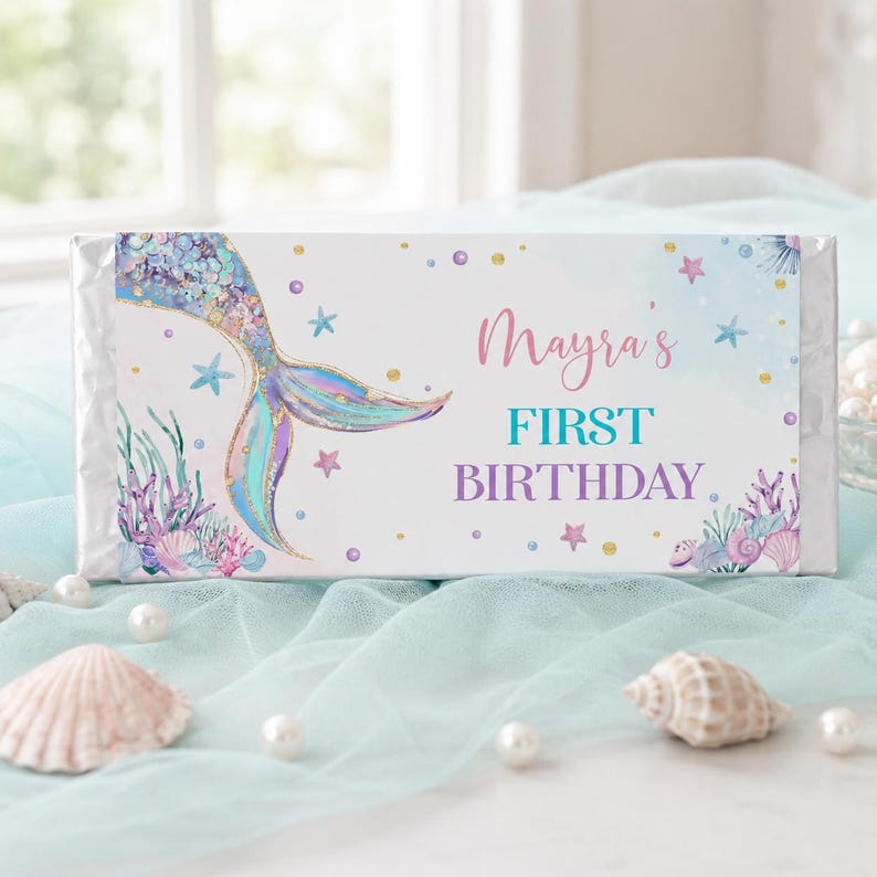 Op de afbeelding: Een rechthoekige wikkel voor een chocoladereep met een illustratie van een zeemeerminstaart, koraal, schelpen en kleine sterretjes. De tekst luidt Mayra's FIRST BIRTHDAY in roze, blauwgroene en paarse letters op een witte achtergrond met gouden stippen.