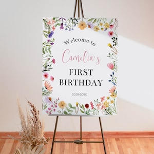 K&ouml;nnte beinhalten: Ein rechteckiges Schild auf einer Metallstaffelei mit einem Aquarell-Blumenrand aus verschiedenen bunten Wildblumen. Der Text lautet Welcome to Camelia's FIRST BIRTHDAY 20.09.202X in schwarzer und rosa Schrift auf wei&szlig;em Grund.