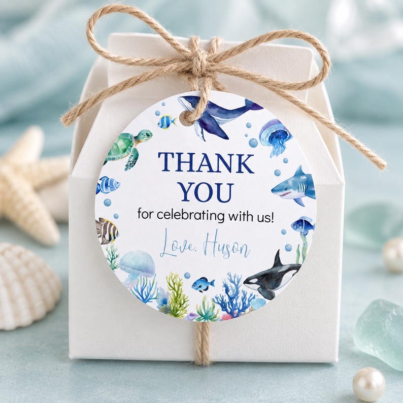 Pu&ograve; includere: Una scatolina quadrata bianca per bomboniere legata con spago naturale e un'etichetta circolare con illustrazioni ad acquerello di creature marine tra cui balena, squalo, tartaruga e medusa, con il testo THANK YOU for celebrating with us! Love, Huson.