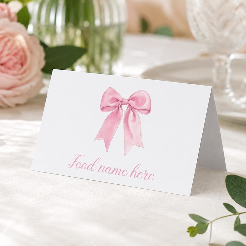 May include: A white folded card featuring a watercolor illustration of a pink ribbon bow centered above the light pink text that reads Food name here. The card is designed to stand upright on a flat surface for display purposes.