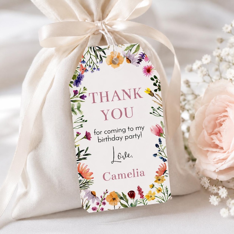 May include: A white fabric drawstring favor bag with a satin ribbon tie, featuring a rectangular paper gift tag. The tag has a floral border with colorful watercolor flowers and the text: THANK YOU for coming to my birthday party! Love. Camelia.