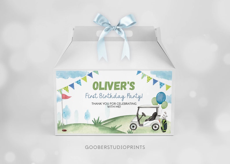 K&ouml;nnte beinhalten: Eine wei&szlig;e Geschenkschachtel mit einem hellblauen Satinband oben. Die Vorderseite zeigt ein Golf-Motiv in Aquarelloptik mit gr&uuml;nen H&uuml;geln, einem Golfcart, Luftballons und Wimpeln. Der Text lautet: OLIVER'S First Birthday Party! THANK YOU FOR CELEBRATING WITH ME!