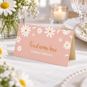 Op de afbeelding: Een gevouwen roze naamkaartje met een wit madeliefjespatroon. De tekst "Food name here" is gedrukt in een goudkleurig scriptlettertype, gevolgd door de tekst "EDITABLE TEMPLATE" in een wit schreefloos lettertype. De kaart staat op een witte ondergrond.