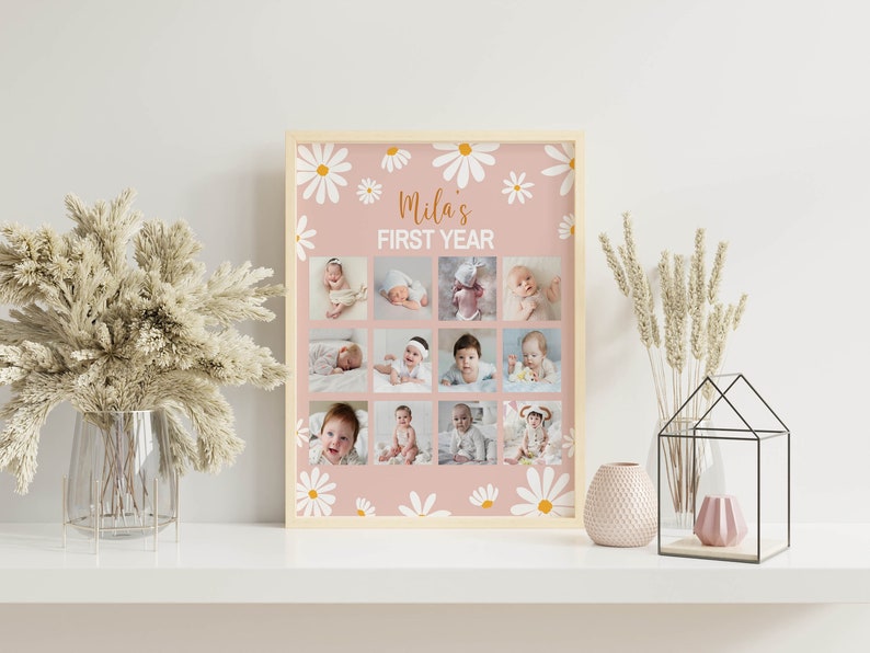 May include: A rectangular photo collage print in a light wood frame featuring a dusty pink background with white daisy illustrations. The text reads Mila's FIRST YEAR above twelve individual square photographs of a baby at different ages.