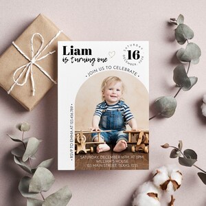 May include: A rectangular birthday invitation card featuring a photo of a toddler in denim overalls and a striped shirt sitting with wooden toy airplanes. The text reads: Liam is turning one, Saturday December 16, 202X, Join us to celebrate, RSVP to Emma at 123.456.789, Saturday, December 16th, at 2pm, William's house, 123 Main Street, Texas. 1235.