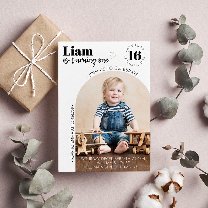 May include: A rectangular birthday invitation card featuring a photo of a toddler in denim overalls and a striped shirt sitting with wooden toy airplanes. The text reads: Liam is turning one, Saturday December 16, 202X, Join us to celebrate, RSVP to Emma at 123.456.789, Saturday, December 16th, at 2pm, William's house, 123 Main Street, Texas. 1235.