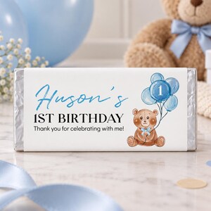 May include: A rectangular candy bar wrapper with a white background featuring a brown teddy bear holding blue balloons. The text reads Huson's 1ST BIRTHDAY Thank you for celebrating with me! The design includes a blue number one on a balloon.