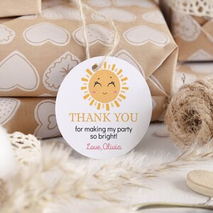 May include: A round white paper gift tag featuring a yellow sun illustration with a smiling face and pink cheeks. The text reads THANK YOU for making my party so bright! Love, Olivia. The tag is attached to a piece of light brown twine string.