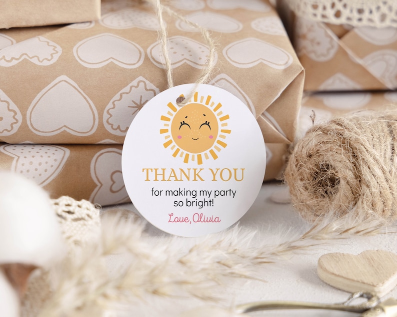 May include: A round white paper gift tag featuring a yellow sun illustration with a smiling face and pink cheeks. The text reads THANK YOU for making my party so bright! Love, Olivia. The tag is attached to a piece of light brown twine string.