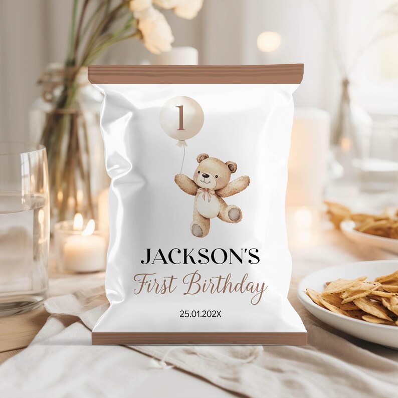 May include: A white chip bag with a brown top. The bag has a design of a teddy bear holding a balloon with the number 1 on it. The text "JACKSON'S First Birthday 25.01.202X" is printed on the bag.