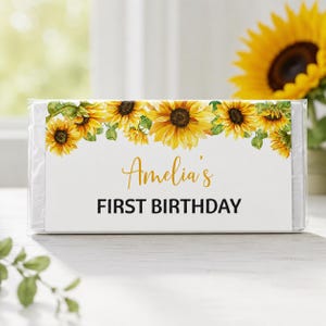 May include: A rectangular candy bar wrapper featuring a white background with a border of yellow sunflowers and green leaves along the top. The text reads Amelia's in a gold script font and FIRST BIRTHDAY in bold black capital letters.