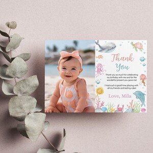 May include: A rectangular thank you card featuring a photograph of a smiling baby on a beach on the left side. The right side displays watercolor illustrations of sea creatures and the text: Thank You, Thank you so much for celebrating my birthday with me and for the wonderful present you gave me! I had such a good time playing with all my friends and eating cake! Love, Mila.