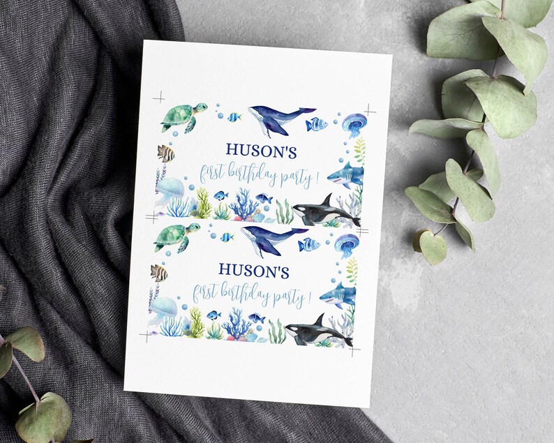 May include: A white rectangular card featuring two identical prints of an underwater scene with watercolor illustrations of a sea turtle, whale, shark, orca, and jellyfish. The text HUSON'S first birthday party! is printed in blue ink.