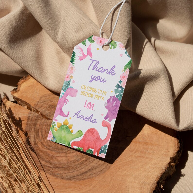 K&ouml;nnte beinhalten: Ein rechteckiges wei&szlig;es Geschenkanh&auml;nger-Schild mit Loch und wei&szlig;er Schnur, verziert mit Aquarellillustrationen von rosa, gr&uuml;nen und violetten Dinosauriern, tropischen Bl&auml;ttern und rosa Blumen. Der Text lautet: Thank you FOR COMING TO MY BIRTHDAY PARTY! Love, Amelia.