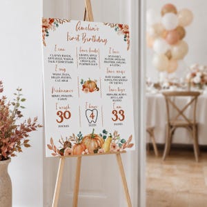 May include: A white rectangular birthday milestone sign on a wooden easel featuring orange pumpkin illustrations and floral accents. The text includes Amelia's First Birthday, I can, Fave foods, I love, I say, Fave songs, Nicknames, Fave toys, I weigh 30 pounds, I have 4 teeth, and I am 33 inches.