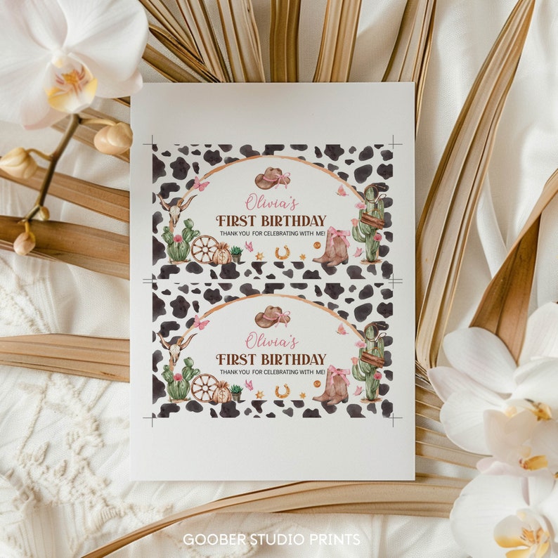 May include: A rectangular card featuring two identical birthday tag designs with a black and white cow print background. The design includes a brown cowboy hat, a skull, a cactus, a wagon wheel, a horseshoe, and boots with pink accents.