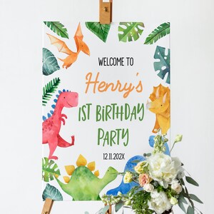 Op de afbeelding: Een rechthoekig bord op een houten ezel met aquarelillustraties van dinosaurussen en tropische bladeren. De tekst luidt: WELCOME TO Henry's 1ST BIRTHDAY PARTY 12.11.202X. Het ontwerp bevat een rode, gele en blauwe dinosaurus met groen blad.