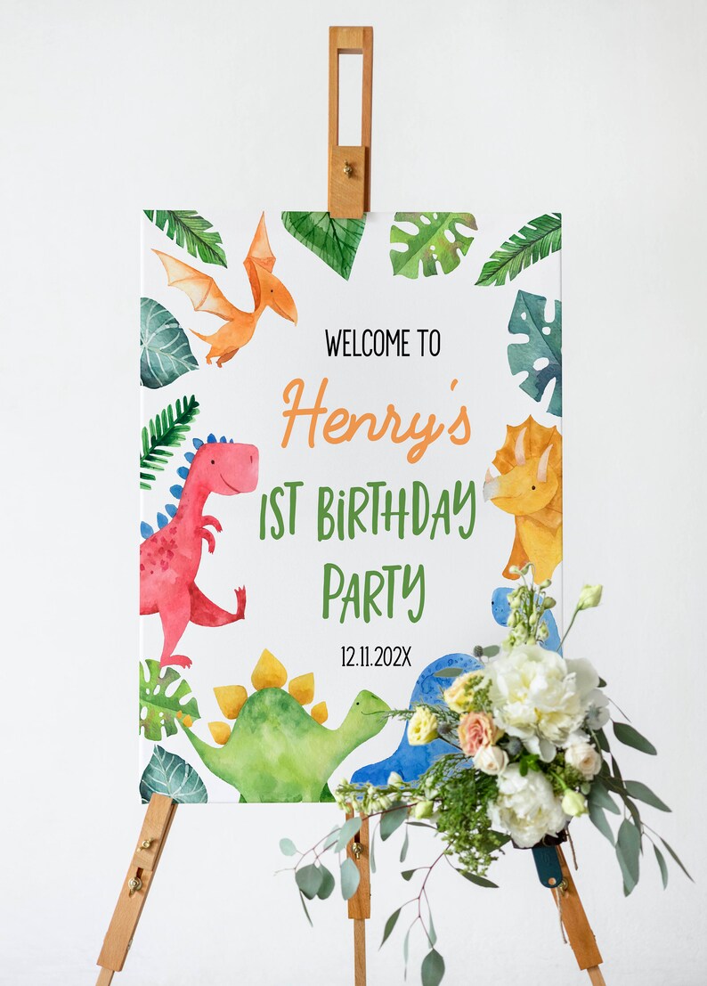 Op de afbeelding: Een rechthoekig bord op een houten ezel met aquarelillustraties van dinosaurussen en tropische bladeren. De tekst luidt: WELCOME TO Henry's 1ST BIRTHDAY PARTY 12.11.202X. Het ontwerp bevat een rode, gele en blauwe dinosaurus met groen blad.