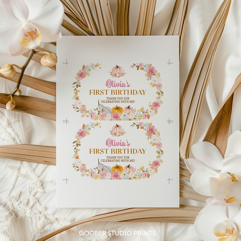 May include: A white rectangular sheet featuring two identical printed labels with floral wreaths in pink, white, and gold. Each label displays the text Olivia's FIRST BIRTHDAY THANK YOU FOR CELEBRATING WITH ME! surrounded by watercolor pumpkins and flowers.