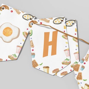May include: A white banner with a breakfast theme. The banner is made of individual flags with a white background and a repeating pattern of breakfast foods like waffles, coffee cups, croissants, and eggs. The flags are connected with a brown string. The word "Happy" is spelled out on the flags in orange letters.