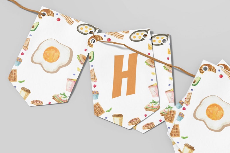 May include: A white banner with a breakfast theme. The banner is made of individual flags with a white background and a repeating pattern of breakfast foods like waffles, coffee cups, croissants, and eggs. The flags are connected with a brown string. The word "Happy" is spelled out on the flags in orange letters.