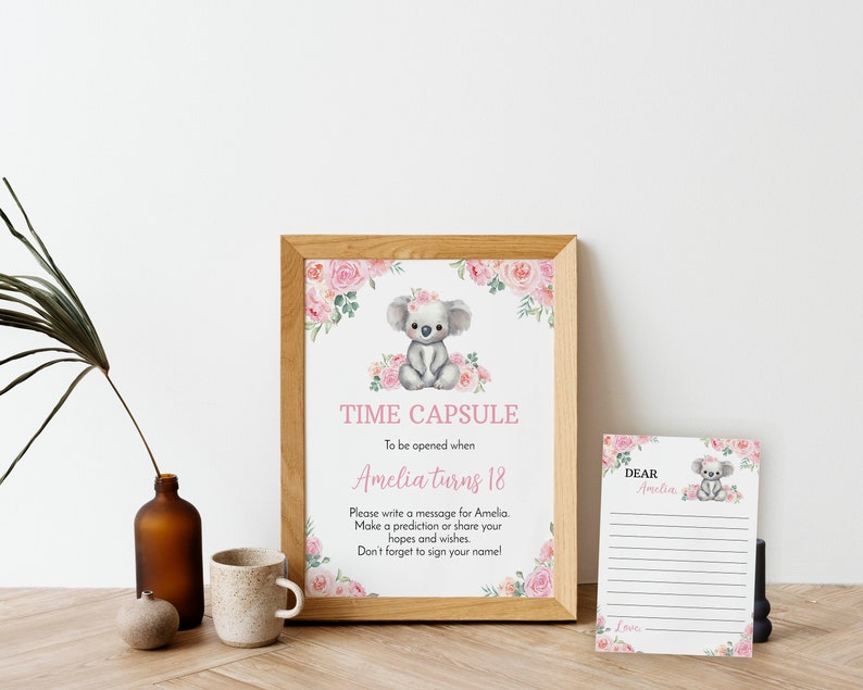 K&ouml;nnte beinhalten: Ein gerahmtes Schild und eine passende Karte mit einer Koala-Illustration und rosa Blumenakzenten. Das Schild tr&auml;gt die Aufschrift TIME CAPSULE To be opened when Amelia turns 18. Please write a message for Amelia. Make a prediction or share your hopes and wishes.