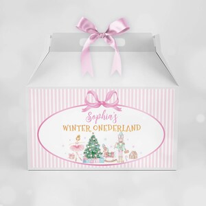 May include: A white gable box with a pink satin ribbon bow on top. The front features a pink striped border and an oval label with the text "Sophia's WINTER ONEDERLAND" above illustrations of a ballerina, a Christmas tree, and a nutcracker.
