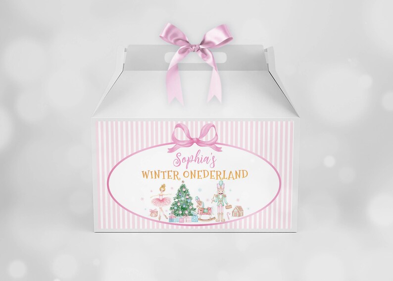 May include: A white gable box with a pink satin ribbon bow on top. The front features a pink striped border and an oval label with the text "Sophia's WINTER ONEDERLAND" above illustrations of a ballerina, a Christmas tree, and a nutcracker.