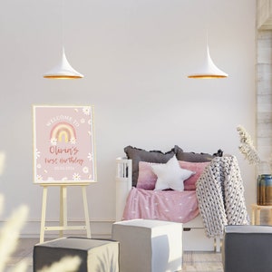 May include: A pink welcome sign with a rainbow and daisies, with the text "Welcome to Olivia's First Birthday 27.02.2023". The sign is on an easel in a room with a white couch and a white star pillow.