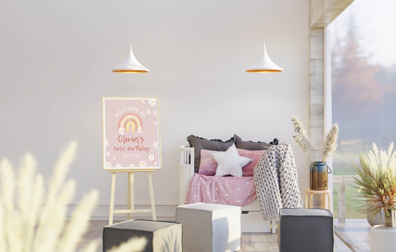 May include: A pink welcome sign with a rainbow and daisies, with the text "Welcome to Olivia's First Birthday 27.02.2023". The sign is on an easel in a room with a white couch and a white star pillow.