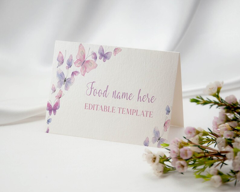 Peut inclure: Une carte blanche pli&eacute;e &agrave; la surface textur&eacute;e orn&eacute;e de papillons &agrave; l'aquarelle dans des tons roses et violets. Le texte "Food name here" est imprim&eacute; en police cursive violette, suivi du texte "EDITABLE TEMPLATE" en police rose sans empattement.