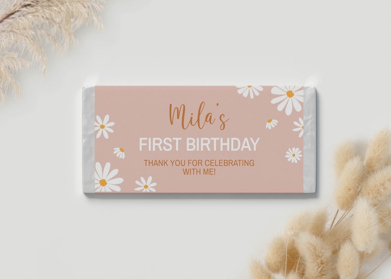 Op de afbeelding: Een roze en witte chocolade reep verpakking met een bloemenpatroon. De tekst op de verpakking luidt "Mila's First Birthday. Thank you for celebrating with me!"