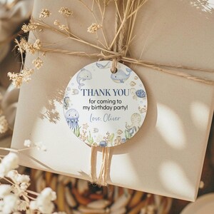 May include: A round gift tag with a marine theme, featuring illustrations of whales, jellyfish, and other sea creatures. The tag reads "THANK YOU for coming to my birthday party! Love, Oliver" in a playful font, tied to a wrapped gift.